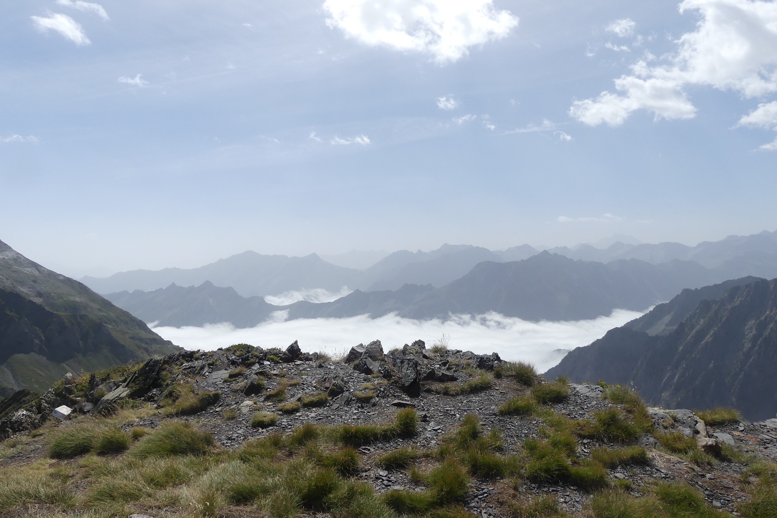 Photo d'une avancée de cailloux et d'herbe qui révèle une mer de nuage et quelques sommets cachés par un voile de nuage